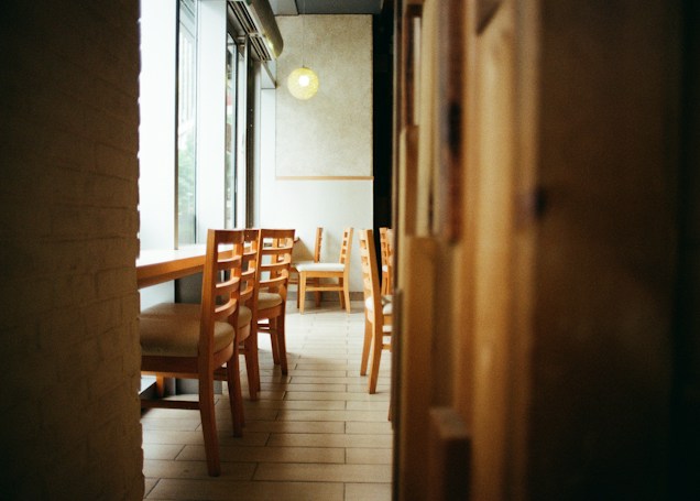 A cozy cafe interior with wooden chairs and tables arranged along a window, allowing natural light to stream in. The warm tones of the wood and soft lighting create an inviting atmosphere.
